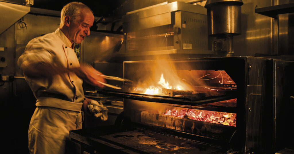 Chef grilling steaks over open flames in the F.X. Buckley kitchen